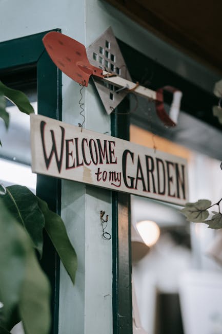 A close-up view of a decorative garden sign hanging from a wooden hook on a blue-green wall inside a garden centre or nursery. The sign reads 'Welcome to my Garden' in bold, artistic lettering, with a