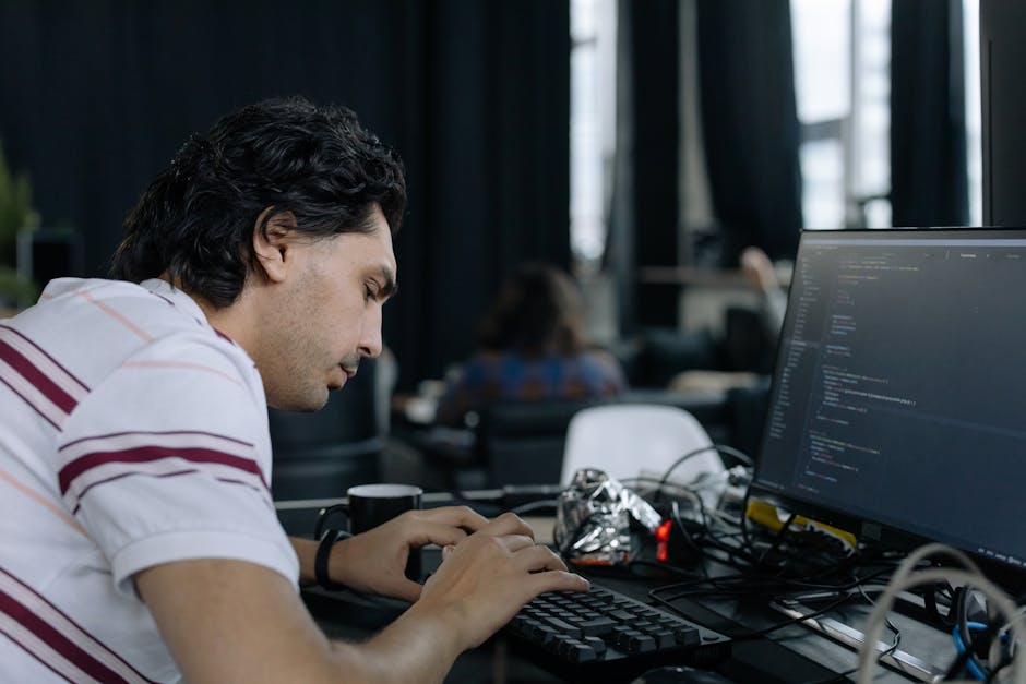 A man with dark, curly hair and light skin is seated at a desk in an indoor workspace, focused on typing on a keyboard. He is wearing a white polo shirt with red and purple horizontal stripes and appe